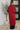 Person wearing a red long coat standing in a room with a plant and framed picture on the wall.