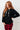Woman wearing a black textured hoodie and red plaid pants against a white wall.