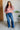 Woman wearing a floral blouse and blue jeans standing indoors against a white brick wall.
