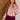 A woman with long wavy red hair models a pink and beige floral blouse with a ruffled front placket, paired with burgundy pants, standing in front of a split wood-paneled and white brick wall background.