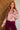 A woman with long wavy red hair models a pink and beige floral blouse with a ruffled front placket, paired with burgundy pants, standing in front of a split wood-paneled and white brick wall background.
