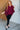 Woman wearing a burgundy top and black pants standing in a room with wooden and white brick walls.