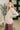 A woman modeling a light beige knit sweater with side slits and a crew neck, paired with mauve pants in a modern indoor setting.