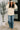 Woman wearing a cream jacket and blue jeans standing indoors next to a plant.