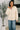 Woman wearing a beige jacket and blue jeans standing indoors with a plant in the background