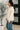 Woman wearing a beige jacket and blue jeans standing indoors with a plant in the background